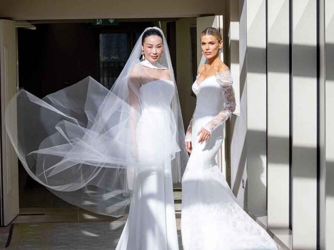 Two brides wearing elegant white wedding gowns stand in a sunlit hallway, one with a flowing veil and the other in a lace-sleeved dress.